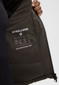 Interno della giacca imbottita Strellson, tessuto verde scuro, tasca con zip, logo bianco del marchio e simboli di cura stampati sulla fodera, texture liscia.