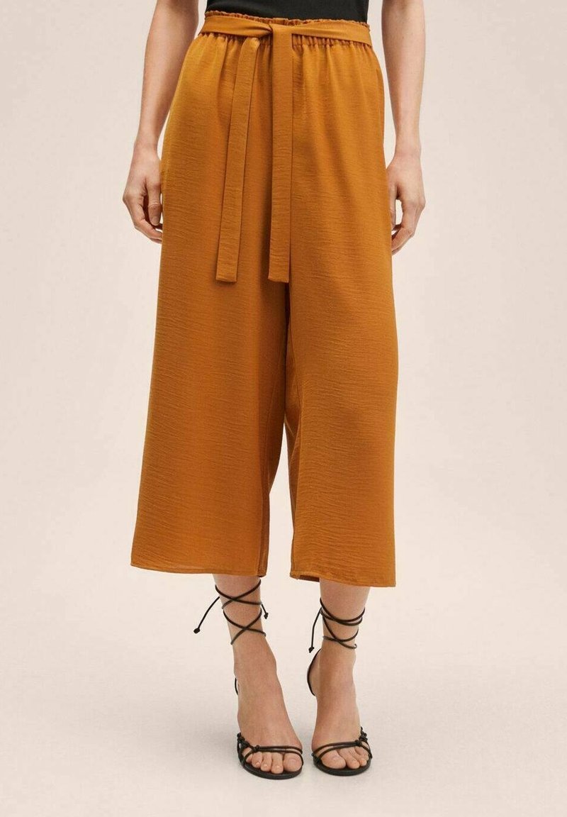 Femme portant un pantalon taille haute à jambes larges couleur rouille avec une ceinture assortie, associé à des sandales à talons noires à brides.