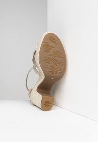 Beżowe sandały na wysokim obcasie z teksturowanym, plecionym wierzchem i gładką, brązową podeszwą z wzorzystym bieżnikiem. Regulowane zapięcie na kostkę.