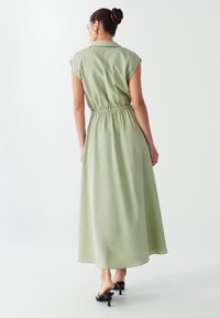 Maxi obleka iz svetlo zelene tkanine, z ovratim, kratkimi rokavi, nabranim pasom in ležernim silhuetom.