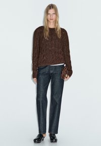 Maglioncino marrone a trecce con maniche lunghe, abbinato a jeans blu a gamba dritta e zoccoli neri, mentre tiene in mano una piccola borsa marrone.