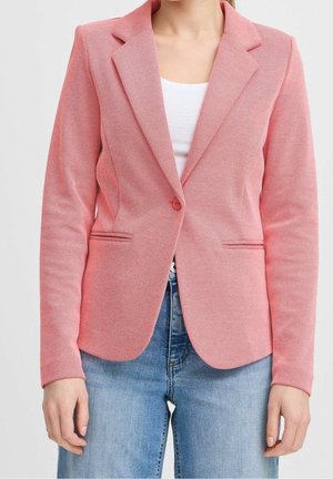 Kvinde iført en tætsiddende lyserød blazer med en enkelt knap, over en hvid top og blå jeans, vist fra nakken til hoften.