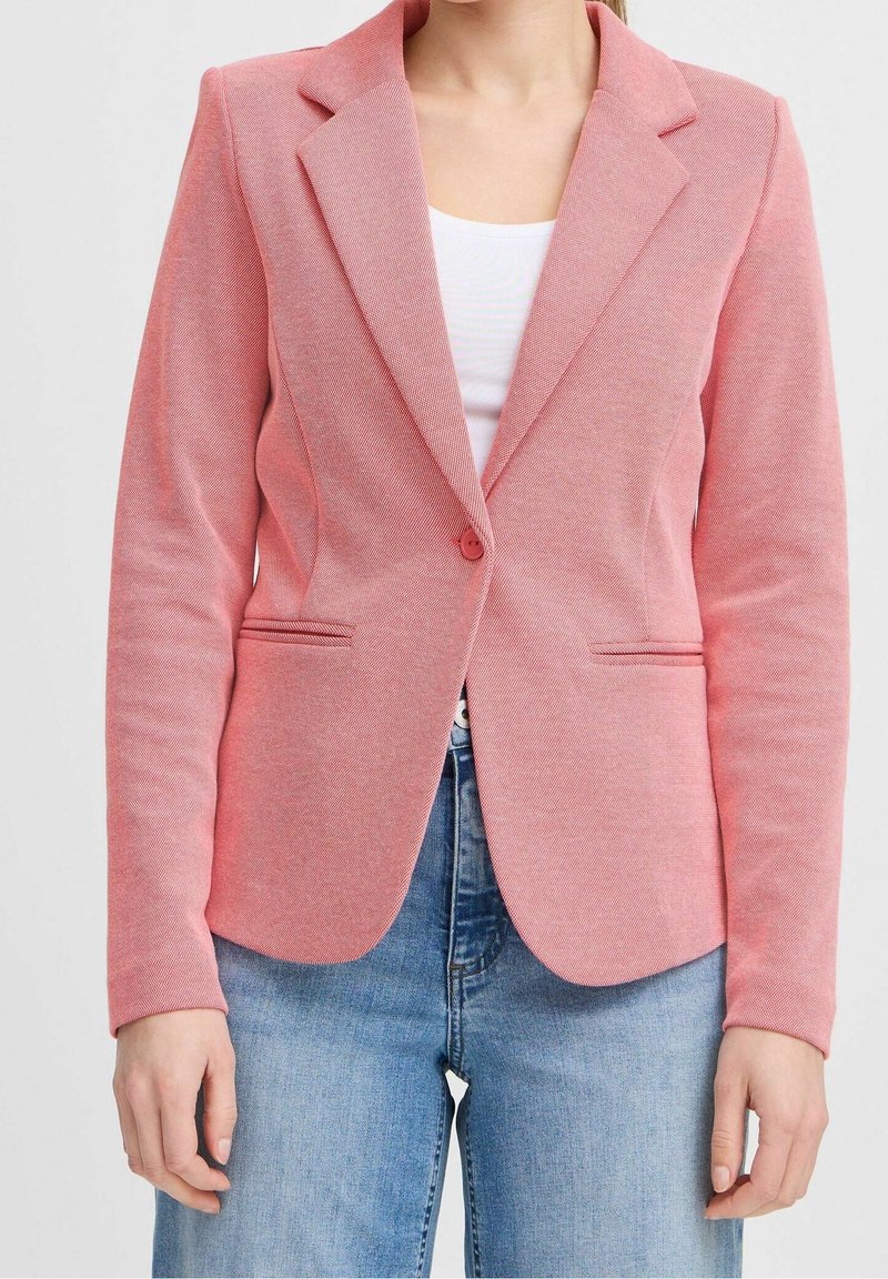 Femme portant un blazer rose ajusté avec un seul bouton, par-dessus un haut blanc et un jean bleu, montrée du cou aux hanches.