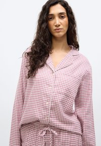 Νεαρή γυναίκα με μακριά σγουρά σκουρόχρωμα μαλλιά, που φοράει ροζ και λευκό καρό πουκάμισο με κουμπιά και παντελόνι που ταιριάζει.