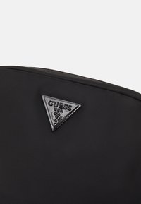 Sort nylon rygsæk med et trekantet sort plastik Guess-logo på fronten. Glat tekstur med et minimalistisk design.