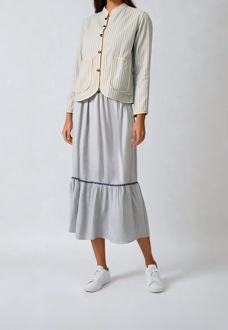 Femme portant une veste légère rayée boutonnée avec un bord jaune et une longue jupe assortie avec un ourlet à volants, associée à des baskets blanches.