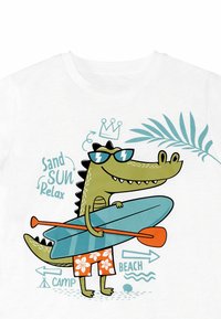 Hvid t-shirt med en tegneserie-krokodille i solbriller, der holder et blåt surfbræt, iført orange og hvide blomstrede shorts, med strand-relateret tekst.