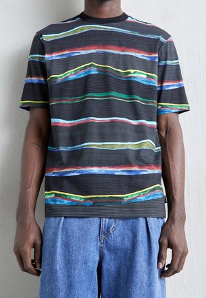Black short-sleeve shirt with colorful irregular horizontal stripes worn by person in blue denim pants against plain wall.