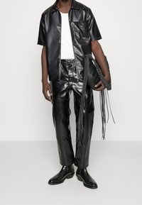 Zwart overhemd van kunstleer met korte mouwen, daaronder een aansluitend wit T-shirt, bijpassende broek en zwarte enkellaarzen. Inclusief een gestructureerde zwarte tas met franjes.