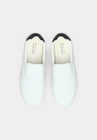 Chaussures blanches sans lacets avec un extérieur texturé, des accents noirs autour de l'ouverture et une doublure intérieure lisse. Présente une étiquette vegan.