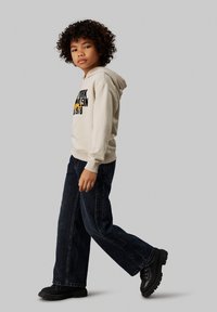 Sudadera beige clara con texto gráfico en negro y amarillo, combinada con jeans de mezclilla oscuros de campana y botas negras gruesas.