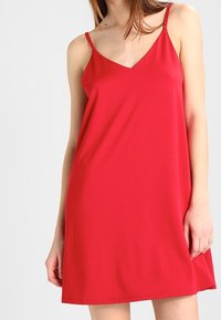 Femme portant une robe rouge sans manches avec des bretelles fines et un décolleté en V sur un fond clair uni.