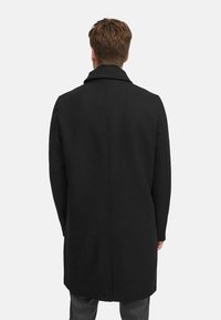 Svart ullfrakk med spiss krage, midtlang design og strømlinjeformet silhuett. Har glatt tekstur og rene sømmer.