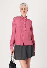 Pink blouse with a high collar, ruffled front, and button closure. Long sleeves with gathered cuffs, worn over a gray pinstriped skirt.
