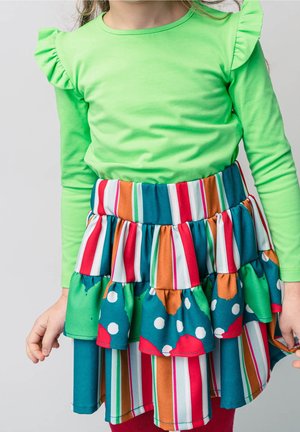 Grünes Langarmshirt mit Rüschendetails, kombiniert mit einem mehrfarbigen Stufenrock mit Streifen und Polka-Dots in Blau, Rot und Grün.