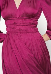 Fuchsia jurk met een V-hals, een geplooide taille en lange mouwen. De stof is glad met een vloeiende, geplooide vormgeving.