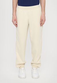 Pantalons de survêtement crème à coupe fuselée, fabriqués dans un tissu doux, dotés d'une taille élastique et de poignets élastiques, associés à des baskets blanches.