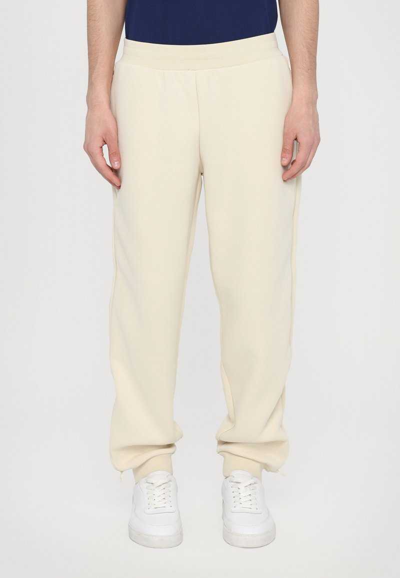 Pantalons de survêtement crème à coupe fuselée, fabriqués dans un tissu doux, dotés d'une taille élastique et de poignets élastiques, associés à des baskets blanches.