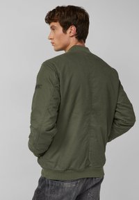 Olivgrüne Bomberjacke mit glatter Textur, gerippten Bündchen und Saum sowie einem dezenten Logo-Patch am linken Ärmel. Rückansicht gezeigt.