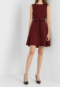 Bordeauxfarbenes ärmelloses Kleid mit tailliertem Oberteil und ausgestelltem Rock, ausgestattet mit einem gebundenen Gürtel in der Taille und einer glatten Stofftextur.