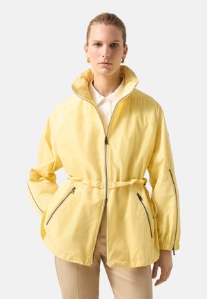 Woman with pulled-back hair wearing gold hoop earrings, a yellow zip-up jacket over a white collared shirt, and beige pants, standing with hands in pockets.