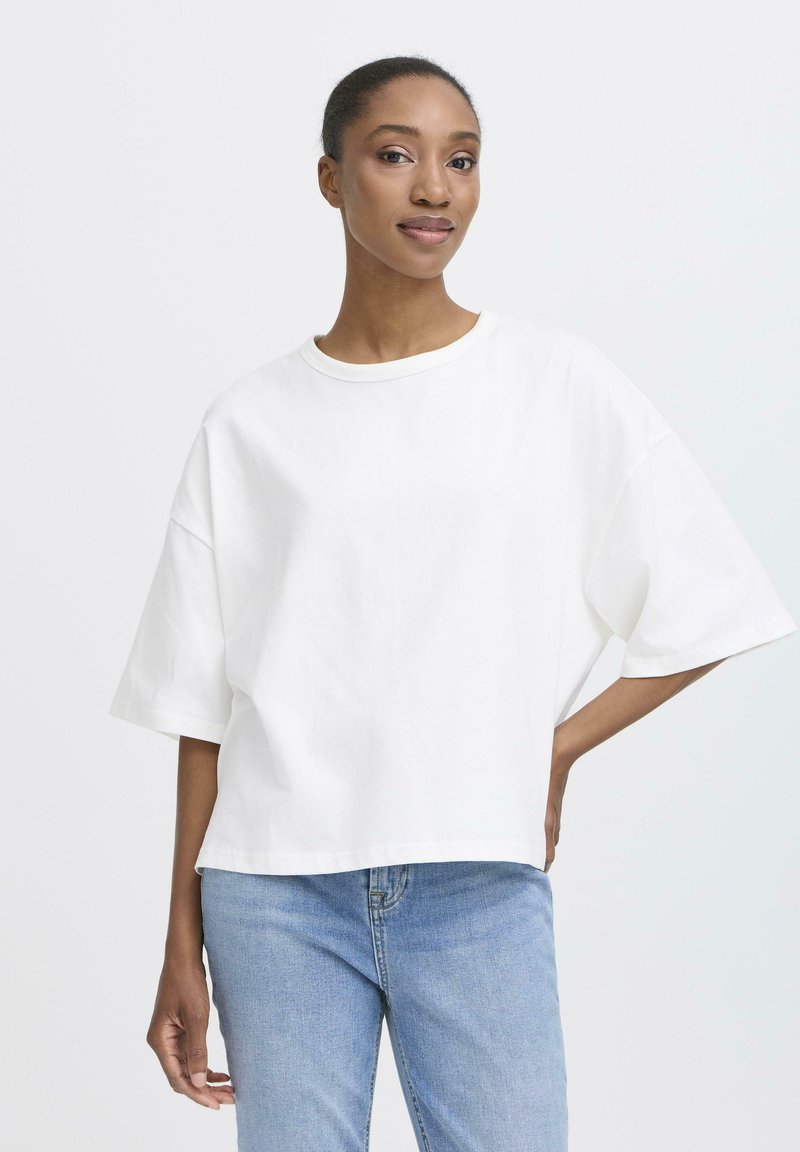 Kvinde med tilbundet hår iført en løs hvid T-shirt og lyseblå jeans, stående foran en ensartet lys baggrund.