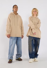 Beige oversized hoodies paired with loose denim jeans. The hoodie has a drawstring; the jeans feature various shades of blue.