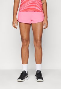 Rosa treningsshorts med en glatt tekstur, kombinert med svarte sko med hvite detaljer og hvite sokker. Toppen har et rødt mønster.
