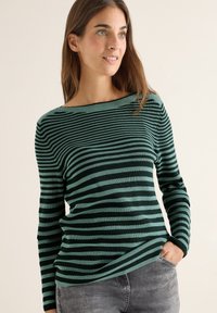 Maglione a maniche lunghe verde e nero a righe con scollo a barca, texture a costine e vestibilità rilassata, abbinato a jeans grigi.
