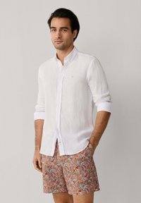 Homme portant une chemise blanche à manches longues retroussées et un short à motif floral, debout devant un fond uni.