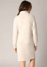 Robe pull en beige clair, avec un col montant, des manches longues et une coupe décontractée. L'ourlet est légèrement au-dessus des genoux. Texture douce.