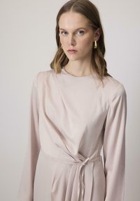Rózsaszín blúzruha hosszú ujjakkal, átkötős dizájnnal, megkötős derék részlettel és sima textúrával. Elegáns, minimalista stílus finom redőzéssel.