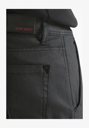 Primer plano de un bolsillo de pantalón negro con costuras cosidas, un lazo para cinturón y una etiqueta rectangular que dice "ANTONY MORATO" en letras rojas.