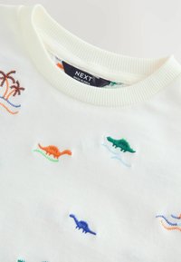 Pull blanc avec des dinosaures colorés brodés en orange, vert, bleu et marron, col rond côtelé et texture douce.