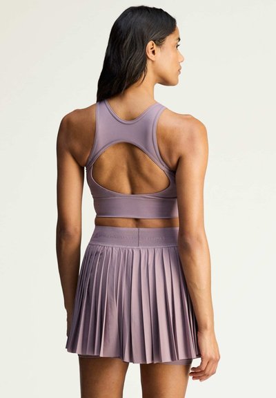 Woman wearing mauve racerback sports bra and high-waisted pleated athletic skirt, standing facing away against plain background.