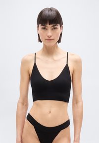 Γυναίκα με κοντά σκούρα μαλλιά φοράει μαύρο ριμπ σουτιέν και εσώρουχο με ψηλό κόψιμο, στέκεται μπροστά σε απλό ανοιχτό φόντο.