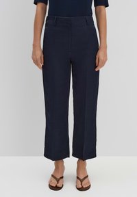 Pantalons en lin bleu marine, taille haute, avec des jambes larges et un devant plat. Présente une texture lisse et un design minimaliste, stylisé avec des sandales.
