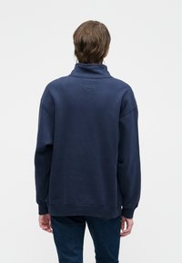 Granatowy sweter z wysokim kołnierzem, luźny krój, ściągacze przy rękawach i u dołu. Gładka tekstura, posiada małą naszywkę z logo z tyłu.