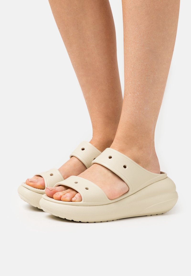 Pieds portant des sandales beiges à enfiler avec deux larges brides et une semelle épaisse et rembourrée sur un fond clair.