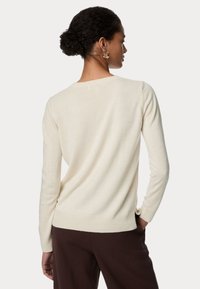 Maglione crema con maniche lunghe, scollatura rotonda e una texture morbida. Indossato con pantaloni marroni, caratterizzati da sottili dettagli a coste all'orlo.