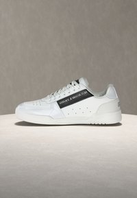 Λευκό sneaker χαμηλού προφίλ με μαύρη ρίγα "Versace Jeans Couture", διάτρητες λεπτομέρειες και κορδόνια στο μπροστινό μέρος, σε ουδέτερο φόντο.