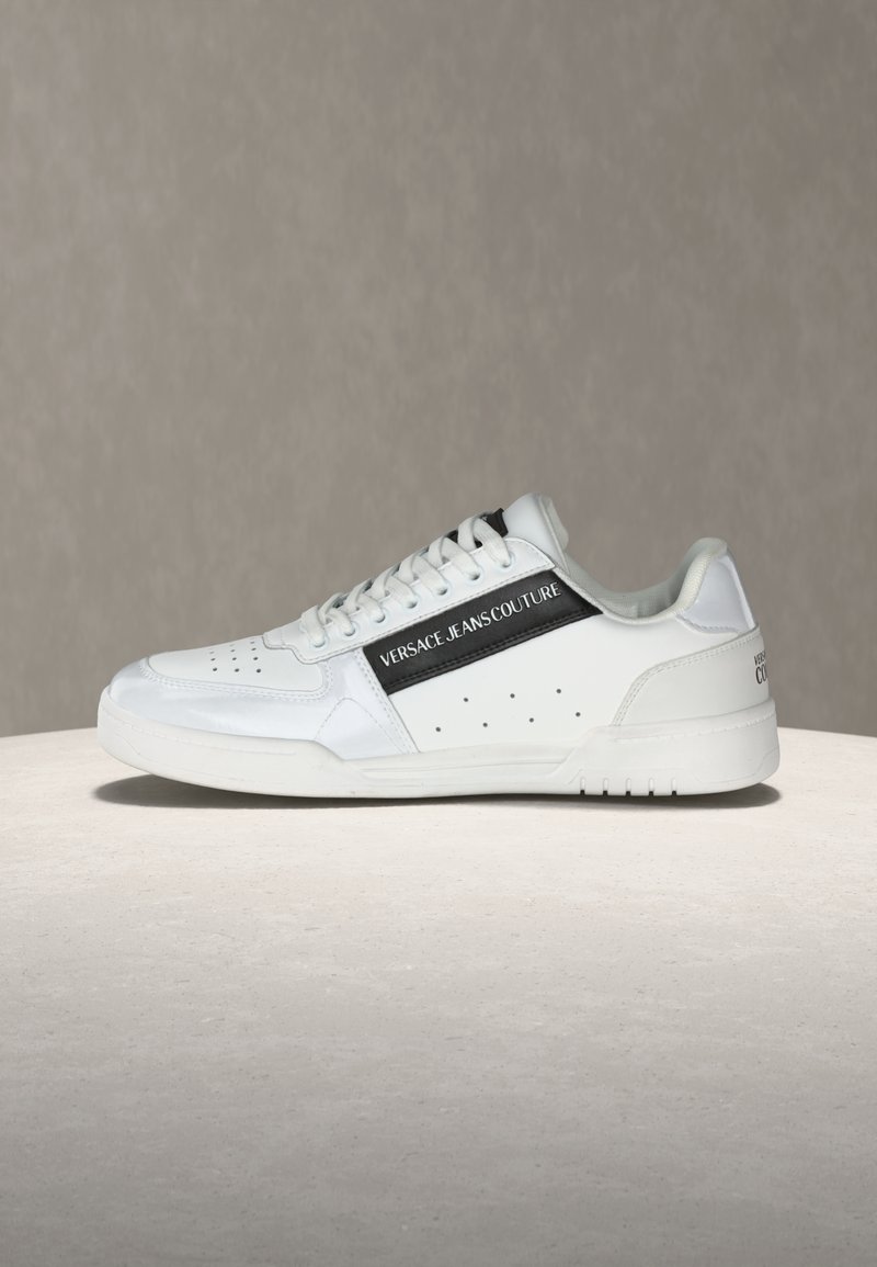 Λευκό sneaker χαμηλού προφίλ με μαύρη ρίγα "Versace Jeans Couture", διάτρητες λεπτομέρειες και κορδόνια στο μπροστινό μέρος, σε ουδέτερο φόντο.