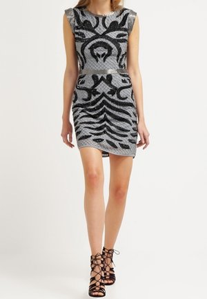 Frau trägt ein silbernes Minikleid mit schwarzen abstrakten Mustern und schwarze, schnürbare High-Heel-Sandalen, steht vor einem weißen Hintergrund.