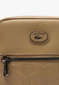 Borsa beige Lacoste con un motivo geometrico testurizzato, chiusura con zip nera e una targa ovale dorata con il logo del coccodrillo.