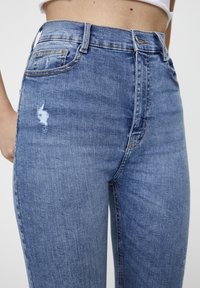 Jeans en denim bleu mi-hauteur avec un léger effet usé et un petit patch déchiré à l'avant, présentant un design classique à cinq poches et des coutures.