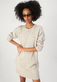 Abito in maglione beige con maniche lunghe, scollatura rotonda e decorato con piccoli strass sul petto. Materiale a maglia morbido e testurizzato.