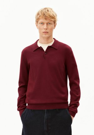 Blonder Mann mit blauen Augen, der ein burgunderfarbenes Langarm-Poloshirt über einem weißen Unterhemd und dunklen Hosen trägt, Hände in den Taschen, neutraler Gesichtsausdruck.