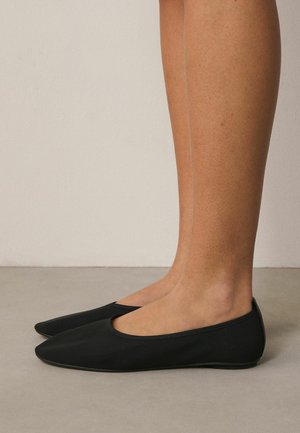 Legs and feet wearing plain black slip-on flats with pointed toes, standing on a smooth beige floor against a light wall background.