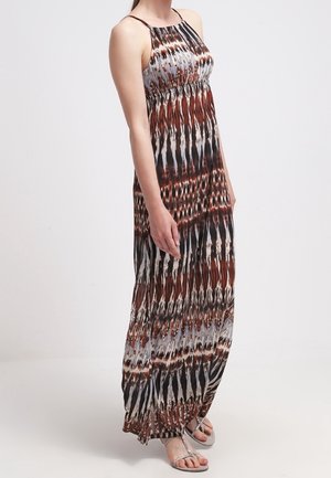 Mujer que lleva un vestido maxi sin mangas y estampado en colores marrón, negro y blanco, combinado con sandalias plateadas sobre un fondo liso.
