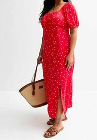 Rotes Kleid mit Punkten, Puffärmeln, einer vorderen Knopfleiste und seitlichem Schlitz. Kombiniert mit braunen Sandalen und einer geflochtenen Tragetasche.